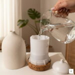 Kobieta nalewa wodę do białego ceramicznego dyfuzora zapachowego PureSense™ na drewnianej podstawie.